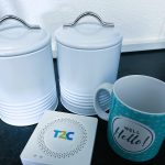 An image of a T2C device which is a white box. It is next to 2 coffee canisters and a coffee mug for scale